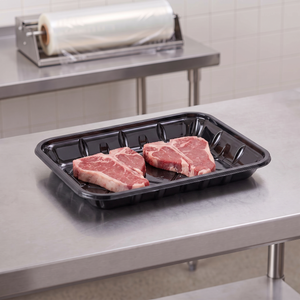 CKF 86515 #4P black PET plastic meat tray holding two raw T-bone steaks on a stainless steel table in a meat packing room