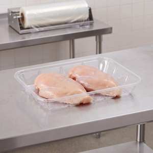 CKF 86605 #4P clear PET plastic meat tray holding two raw boneless chicken breasts on a stainless steel table in a meat packing room