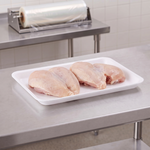 CKF 88116 #16S white foam meat tray holding two raw bone-in chicken breasts on a stainless steel table in a meat packing room