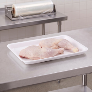 CKF 88149 #9L white foam meat tray holding two raw chicken leg quarters on a stainless steel table in a meat packing room